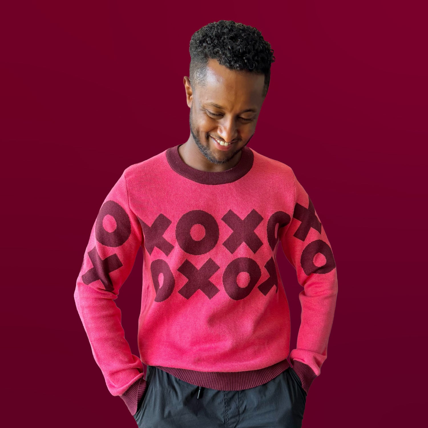 Man wearing a pink sweater with 'XOXO' pattern on a red background