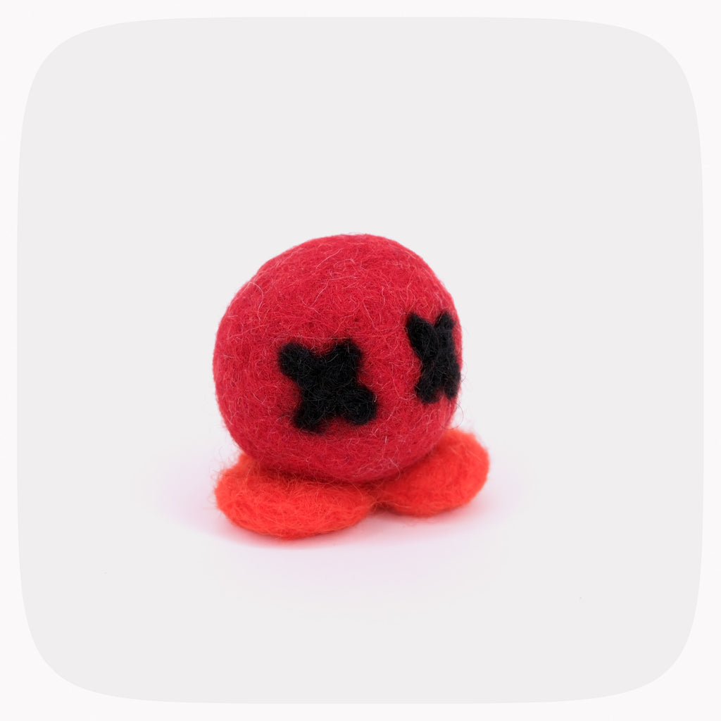 A small felted red ball with black X eyes and orange feet.