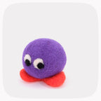 A purple felted ball with eyes looking down and bright orange feet.