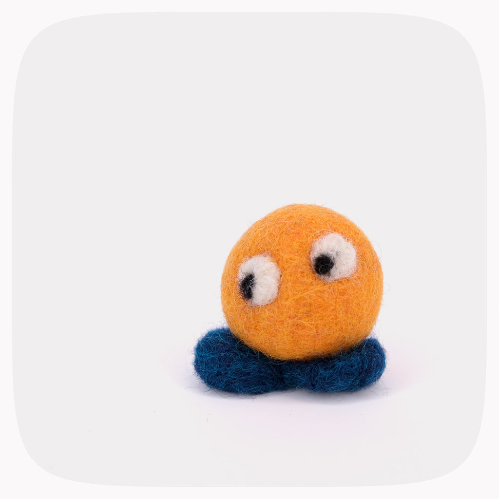 A small yellow felted ball with eyes looking down with blue feet.