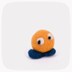 A small yellow felted ball with eyes looking down with blue feet.