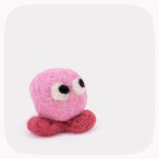 A pink felted ball with eyes looking up and red feet.
