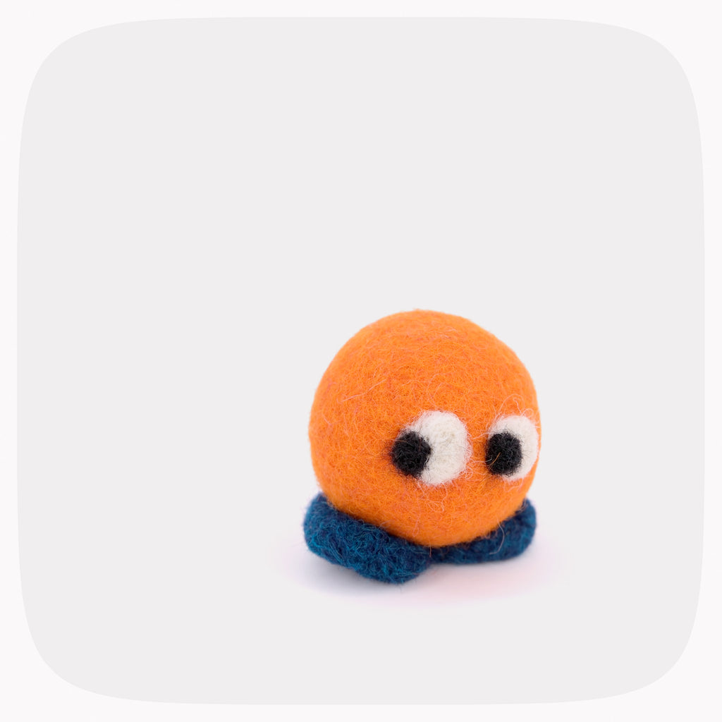 A small yellow felted ball with eyes looking left and blue feet.