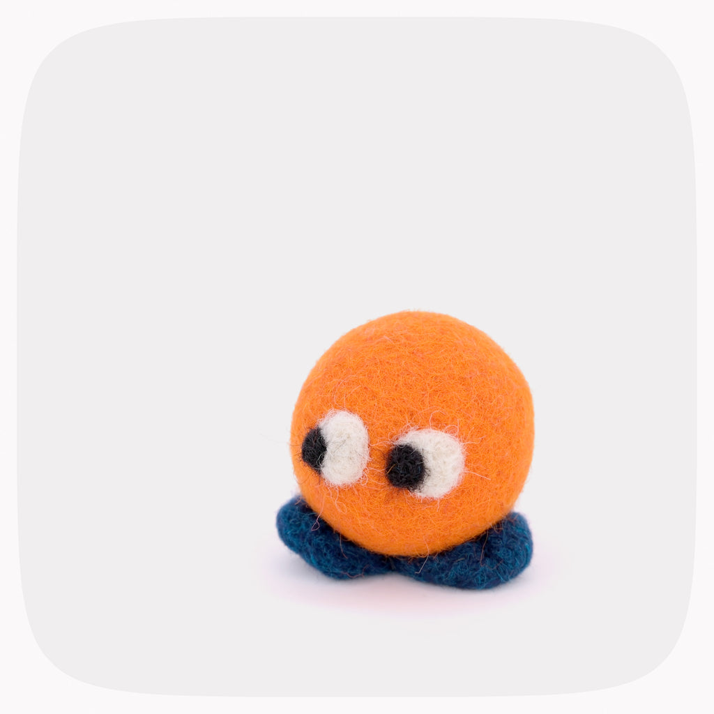 A small yellow felted ball with eyes looking left and blue feet.