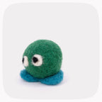 A small felted green ball with blue feet and eyes looking left.
