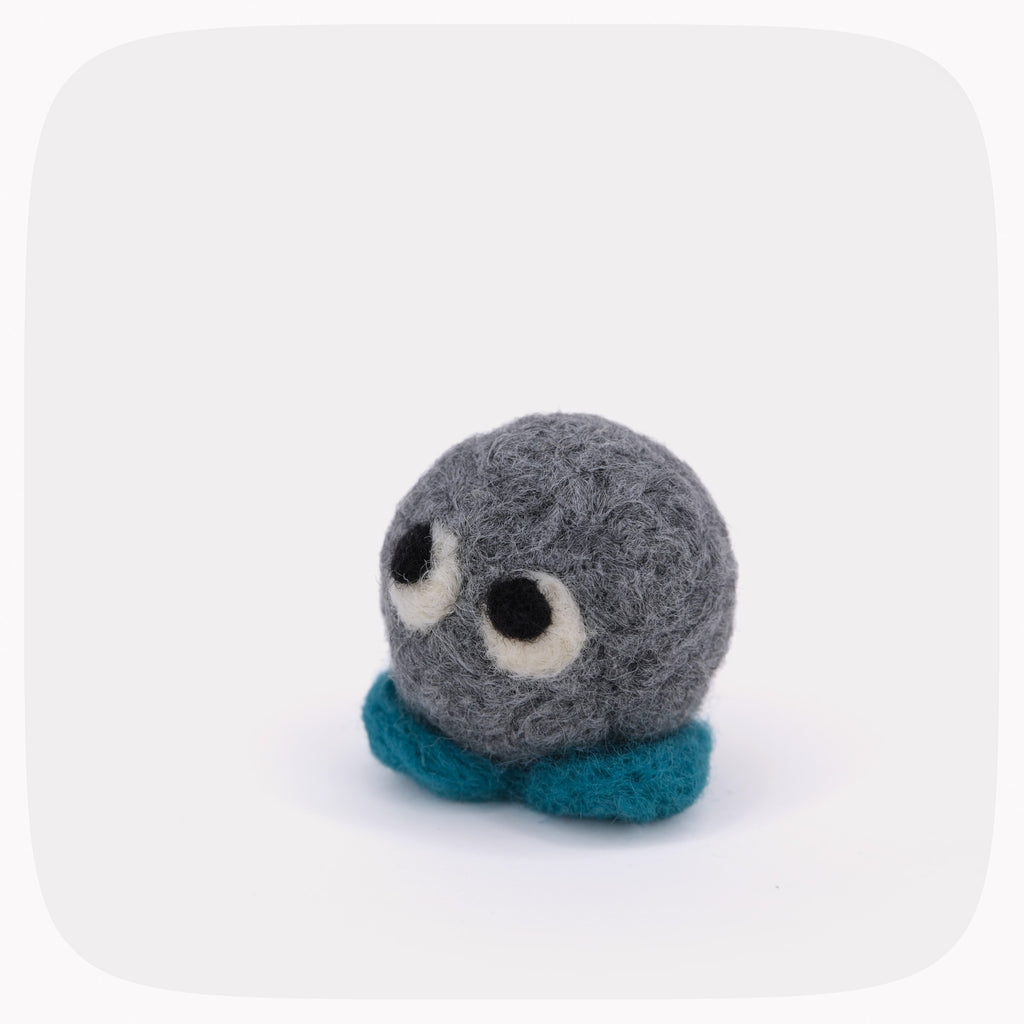 A small handmade felt toy with a round gray head, large white and black eyes, and teal feet, photographed on a clean white background with rounded corners.