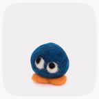 A small felted wool character toy with a round blue body, large white and black eyes, and an orange foot, centered on a light gray rounded square background.