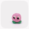 A small felted wool character toy with a round pink head, black and white eyes, and green leaf-like feet, centered on a light gray background with rounded corners.