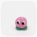 A small felted wool character toy with a round pink head, black and white eyes, and green leaf-like feet, centered on a light gray background with rounded corners.