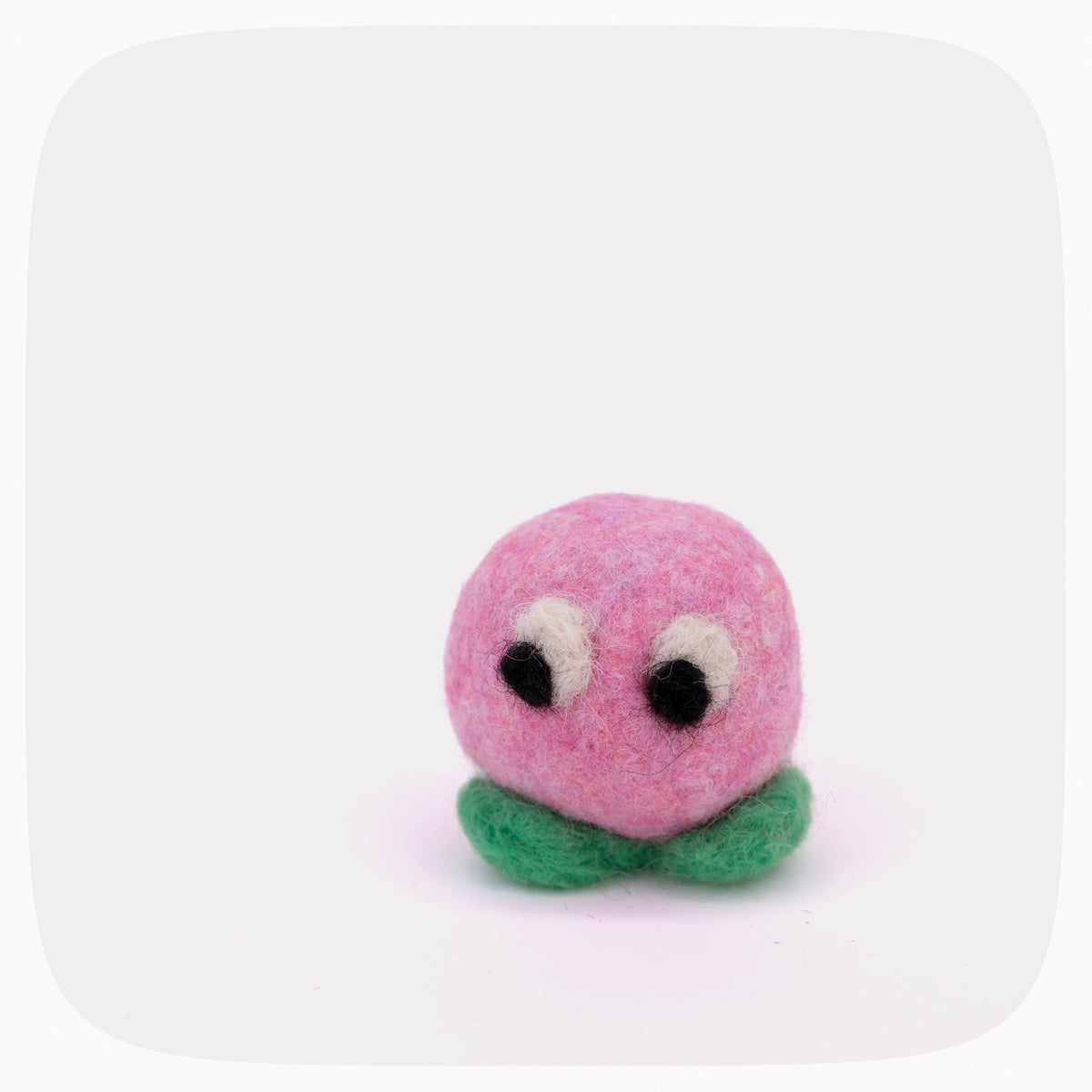 A small felted wool character toy with a round pink head, black and white eyes, and green leaf-like feet, centered on a light gray background with rounded corners.