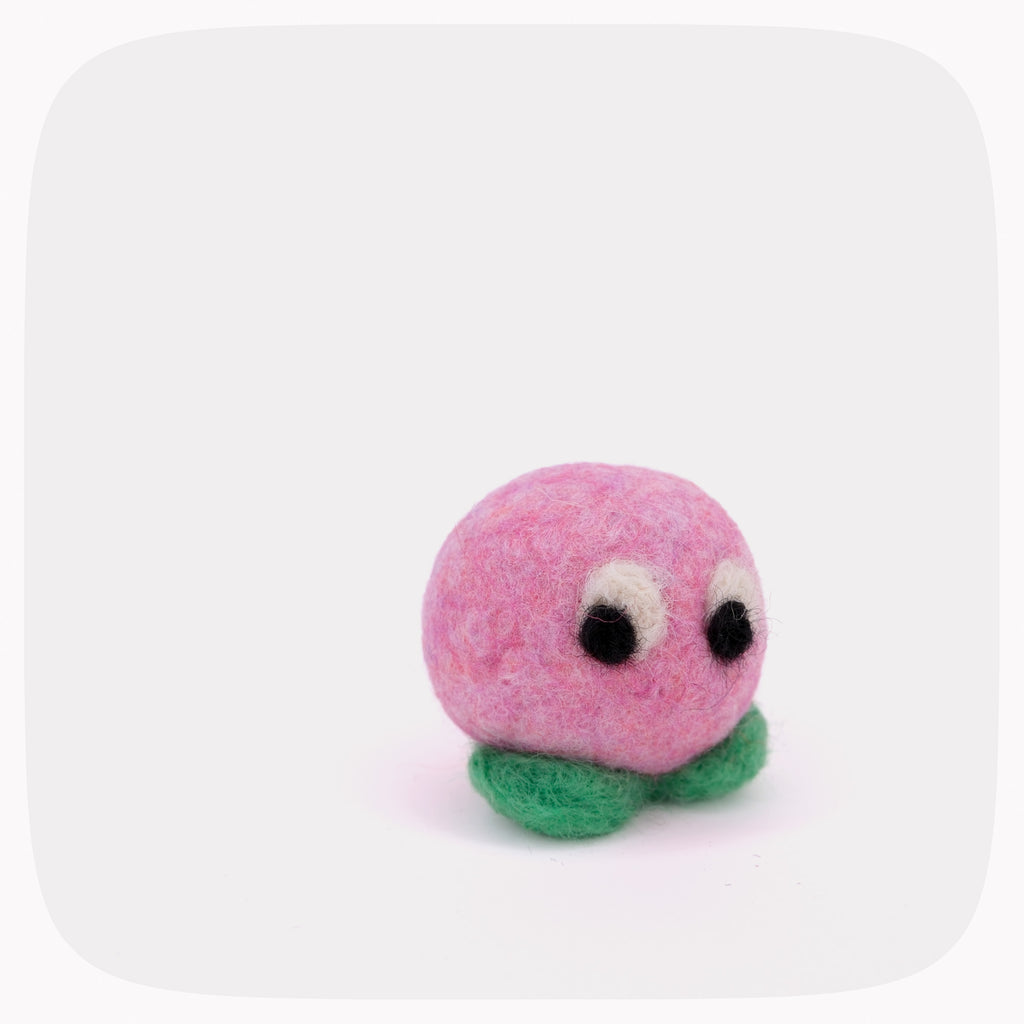 A small pink felt creature toy with a round fuzzy body, green felt feet, and large black-and-white eyes, photographed on a light gray background with rounded corners.