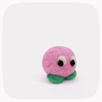 A small pink felt creature toy with a round fuzzy body, green felt feet, and large black-and-white eyes, photographed on a light gray background with rounded corners.