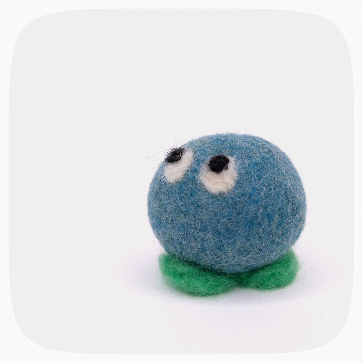A small felted wool creature with a blue round body, green leaf-like base, and two white-and-black eyes, photographed on a light gray background with rounded corners.