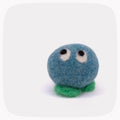A small felted wool creature toy with a round blue body, green feet, and large white eyes with black pupils, centered on a light gray background with rounded corners.