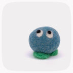 A small felted wool creature toy with a round blue body, green feet, and large white eyes with black pupils, centered on a light gray background with rounded corners.