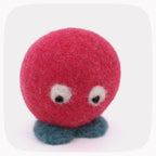 A small felt toy character with a round pink-red head, white eyes with black pupils, and blue feet, photographed on a light gray background with rounded corners.