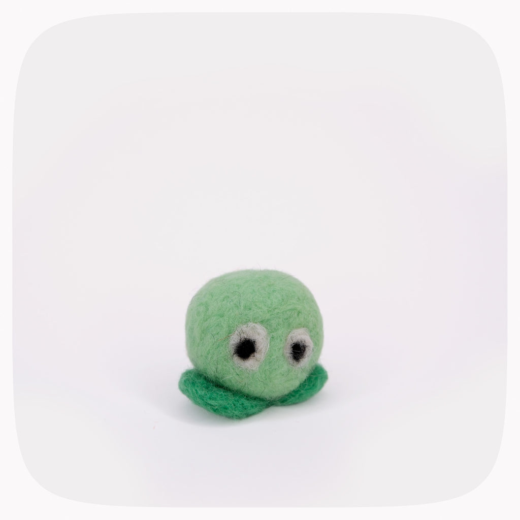 A small green felt toy with a round head, simple black-and-white eyes, and a short curly tail, photographed on a clean white background with rounded corners.