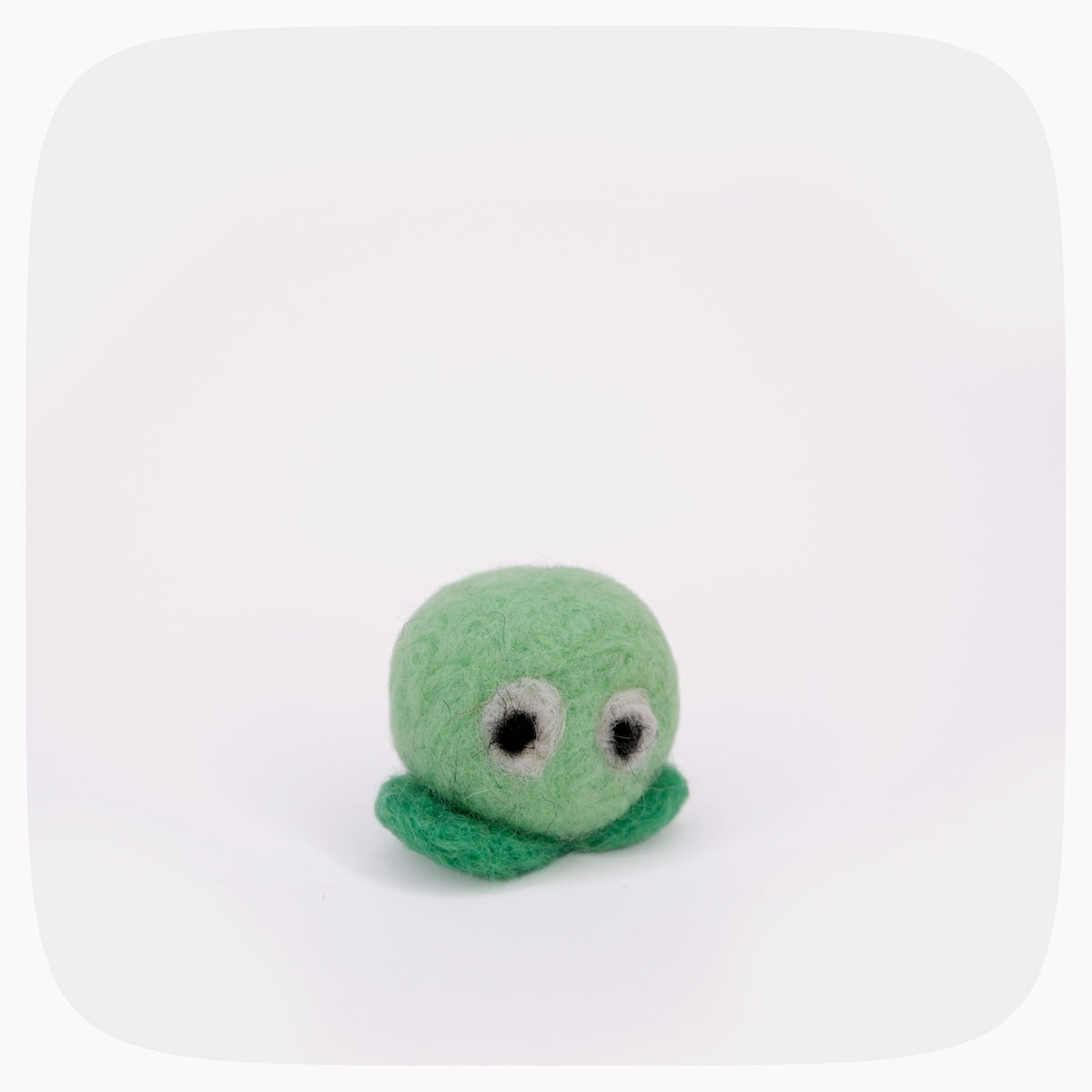 A small green felt toy with a round head, simple black-and-white eyes, and a short curly tail, photographed on a clean white background with rounded corners.