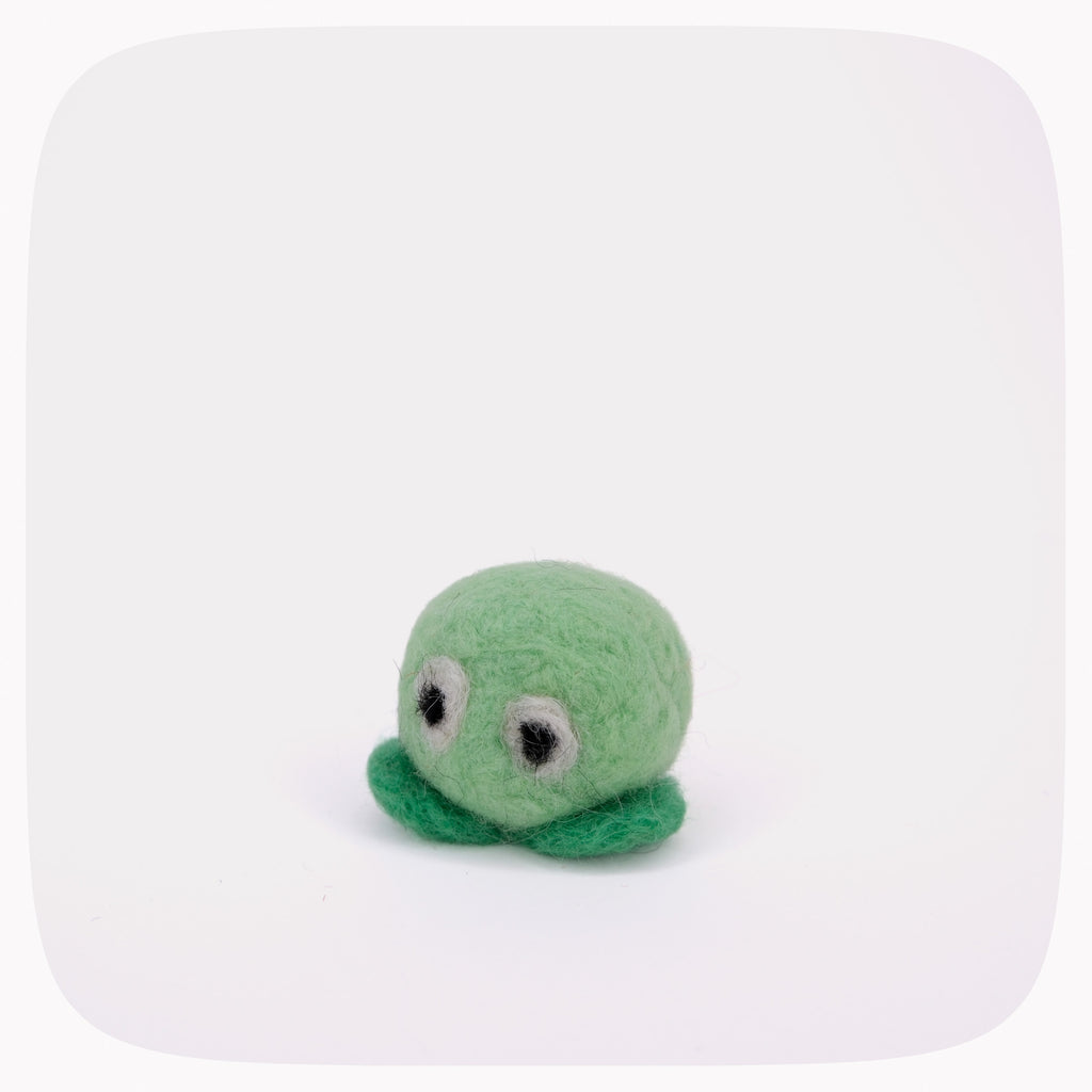 A small green felt toy with large white eyes and dark bead eyes, standing on a dark green felt base against a white background with rounded corners.