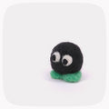A small handmade felt creature with a black round body, large white and black eyes, and a green base, centered on a light gray background with rounded corners.
