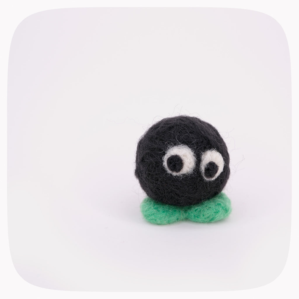 A small handmade felt toy character with a round black body, large white googly eyes, and a mint green base, centered on a white background with rounded corners.