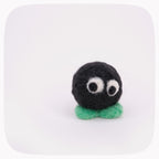 A small handmade felt toy character with a round black body, large white googly eyes, and a mint green base, centered on a white background with rounded corners.