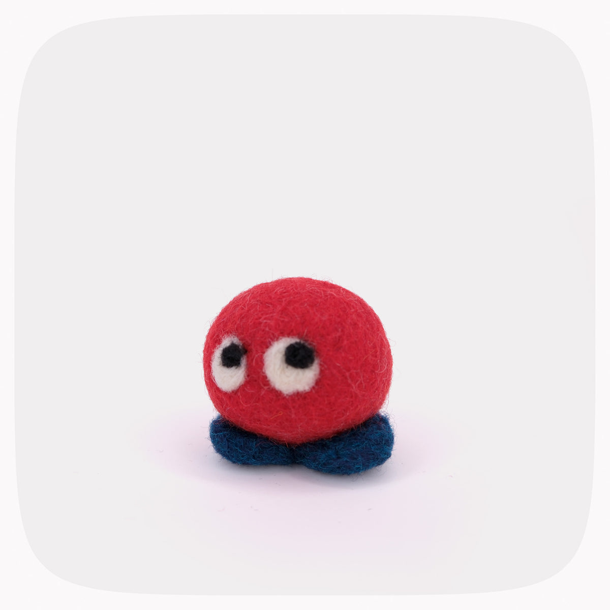 A small felt toy creature with a round red body, large white eyes with black pupils, and two dark blue felt legs, centered on a light gray background.