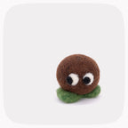 A small felt toy character with a round brown head, white and black eyes, and a green felt base, centered on a light gray background with rounded corners.