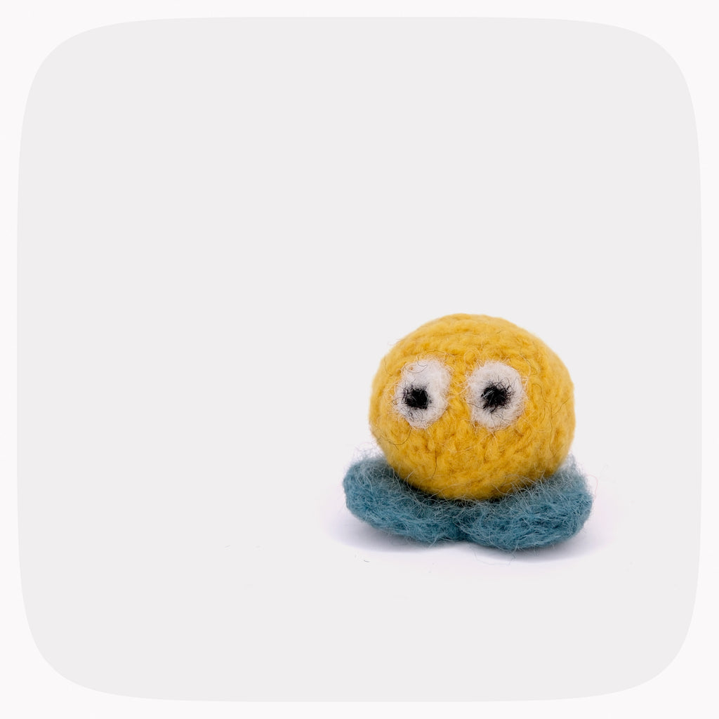 A small handmade felt toy with a round yellow head, googly-style eyes, and a blue-green base, photographed on a plain light gray background.