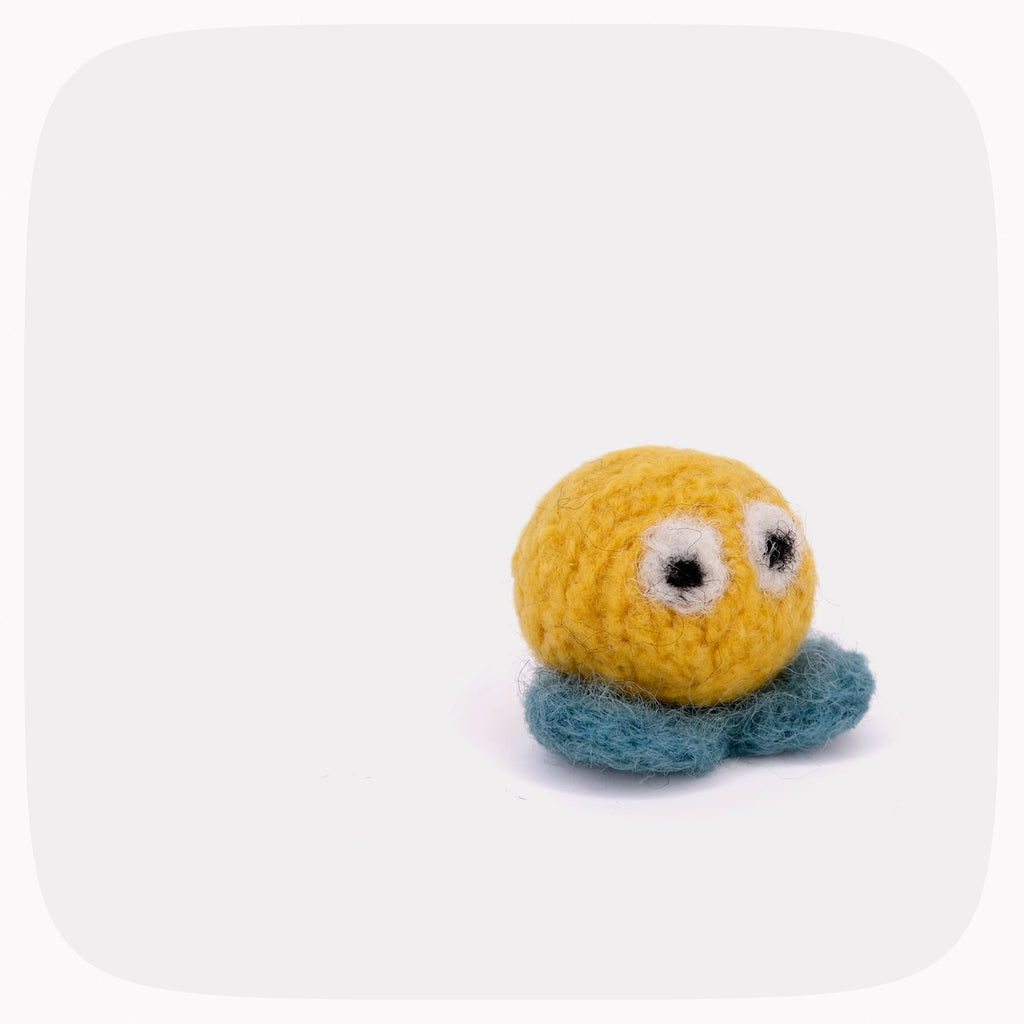 A small handmade felt toy with a round yellow head, blue-green tentacles, and large googly eyes, photographed on a plain light gray background.