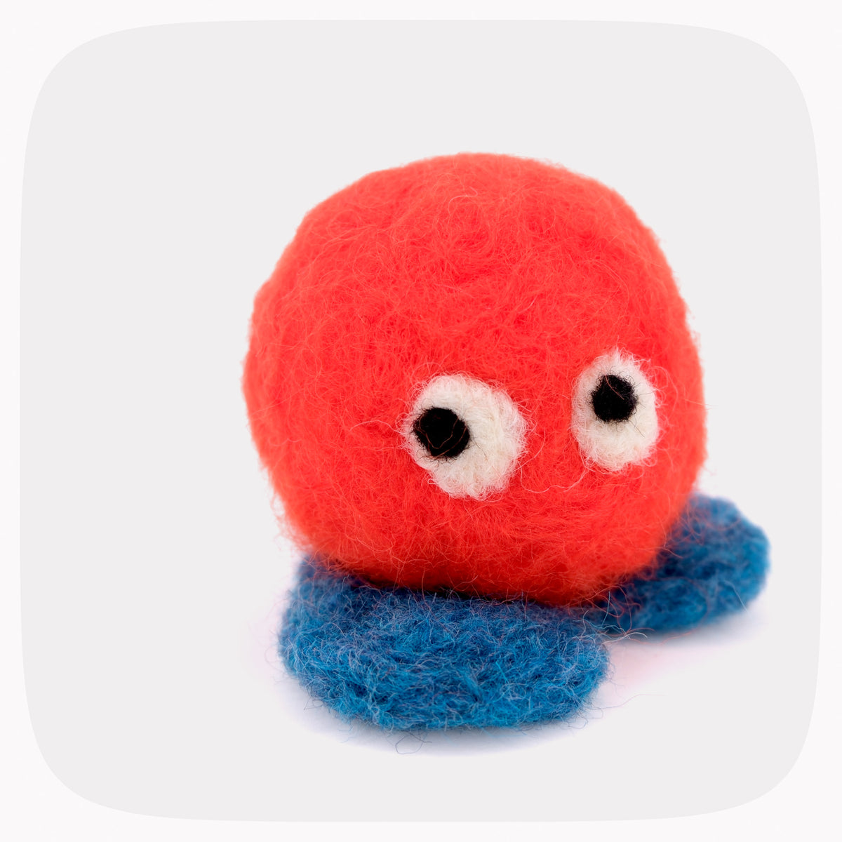 A small felted wool toy with an orange round body, blue feet, and large white eyes with black pupils, photographed on a light gray background with rounded corners.