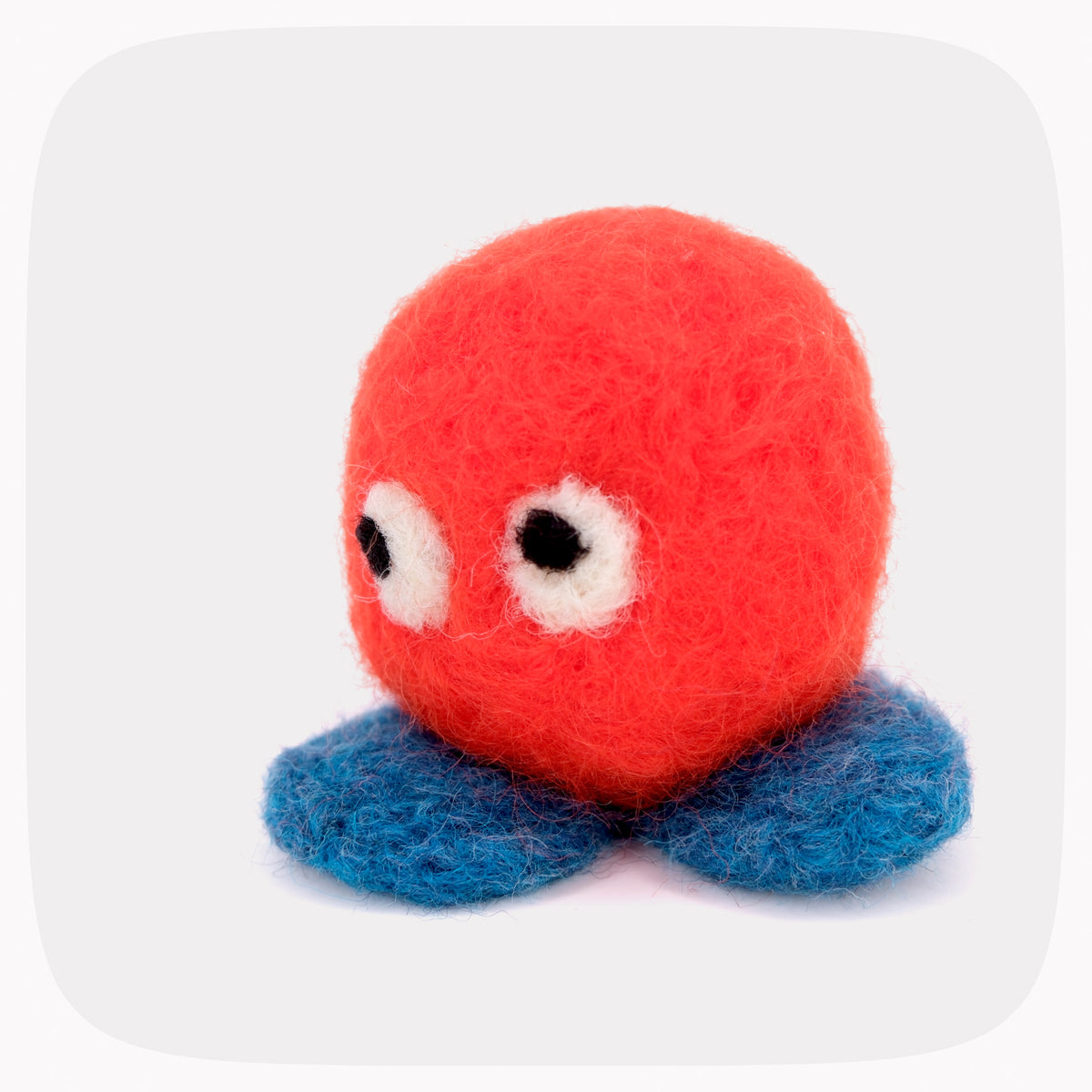 A small felted wool toy with an orange round body, blue feet, and large white eyes with black pupils, photographed on a light gray background with rounded corners.