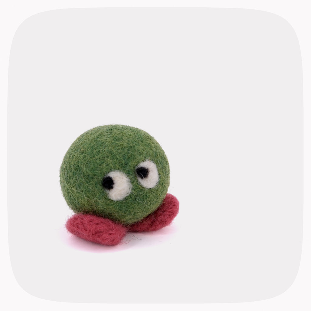 A small felted wool toy with a green round body, red feet, and large white eyes with black pupils, photographed on a light gray background with rounded corners.