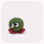A small felted wool toy with a green round body, red feet, and large white eyes with black pupils, photographed on a light gray background with rounded corners.