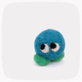 A small felted wool toy character with a round blue head, black and white eyes, and green legs, photographed on a light gray background with rounded corners.