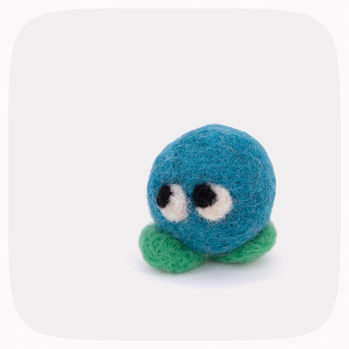 A small felted wool toy with a blue round body, green feet, and large white-and-black eyes, centered on a light gray background with rounded corners.