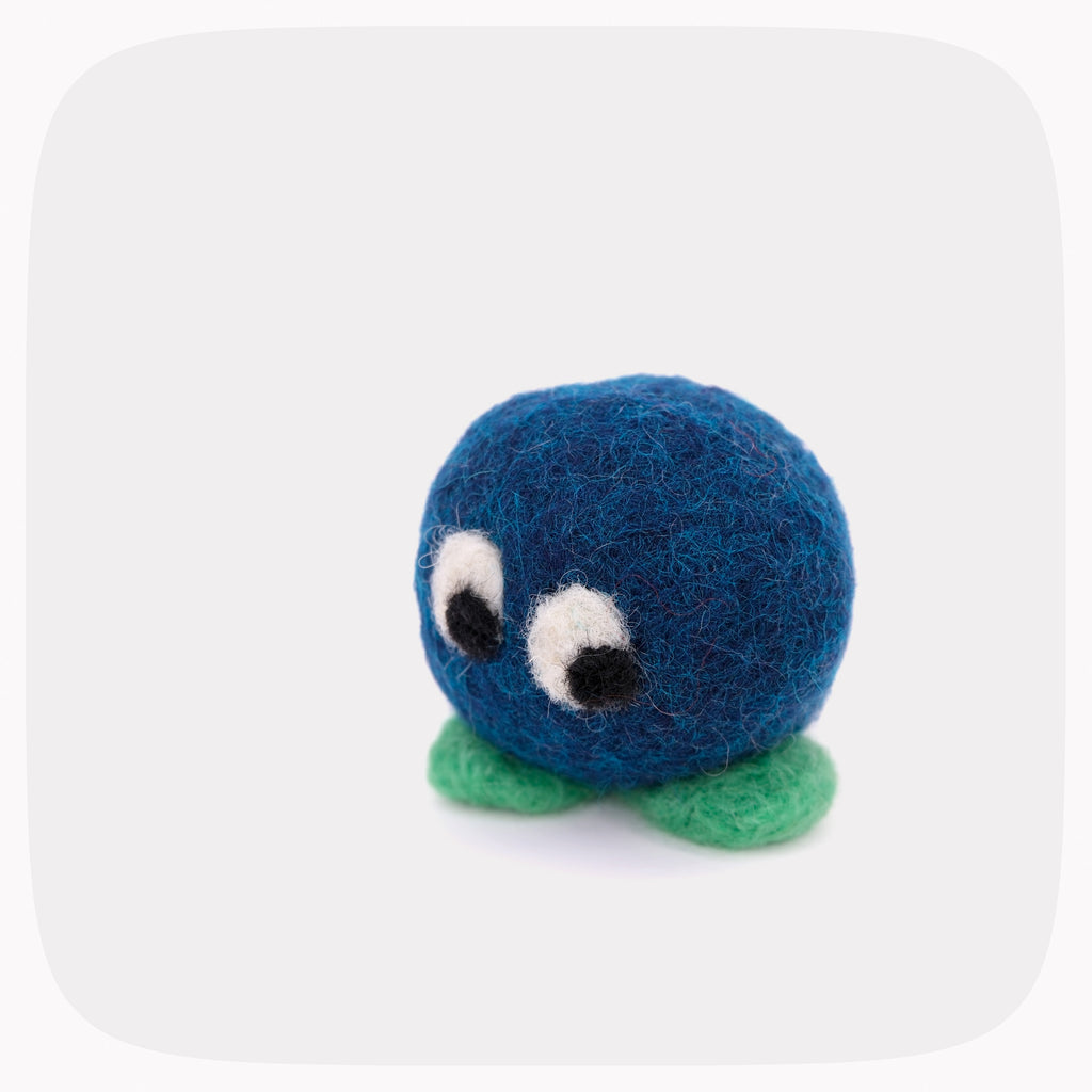 A small blue felt plush toy with large white and black eyes and green felt feet, centered on a light gray rounded-square background.
