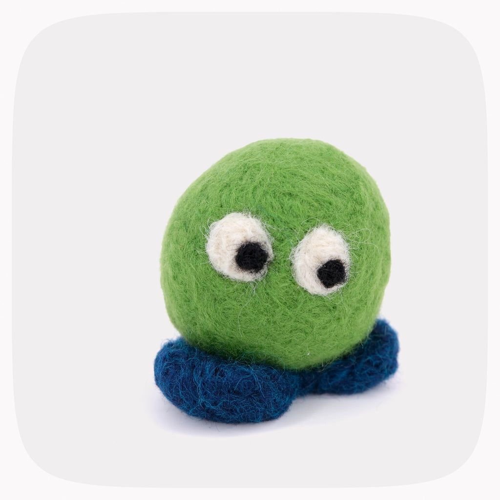 A small felted wool toy with a round green head, two large white eyes with black pupils, and dark blue feet centered on a light gray rounded-square background.