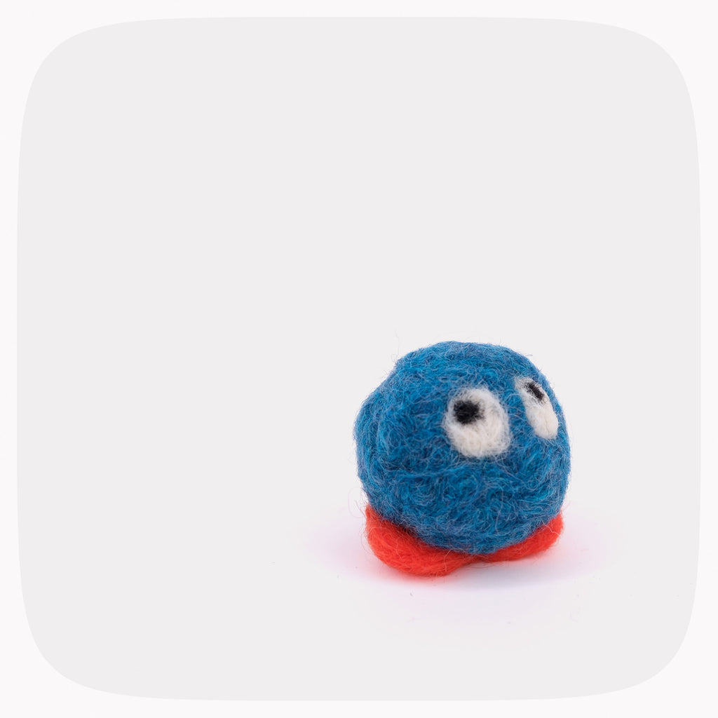 A small felted wool toy character with a blue round body, white eyes, and a red base, photographed on a clean white background with rounded corners.