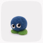 A small felted wool plush toy shaped like a round blue creature with large white eyes and green feet, centered on a light gray background with rounded corners.
