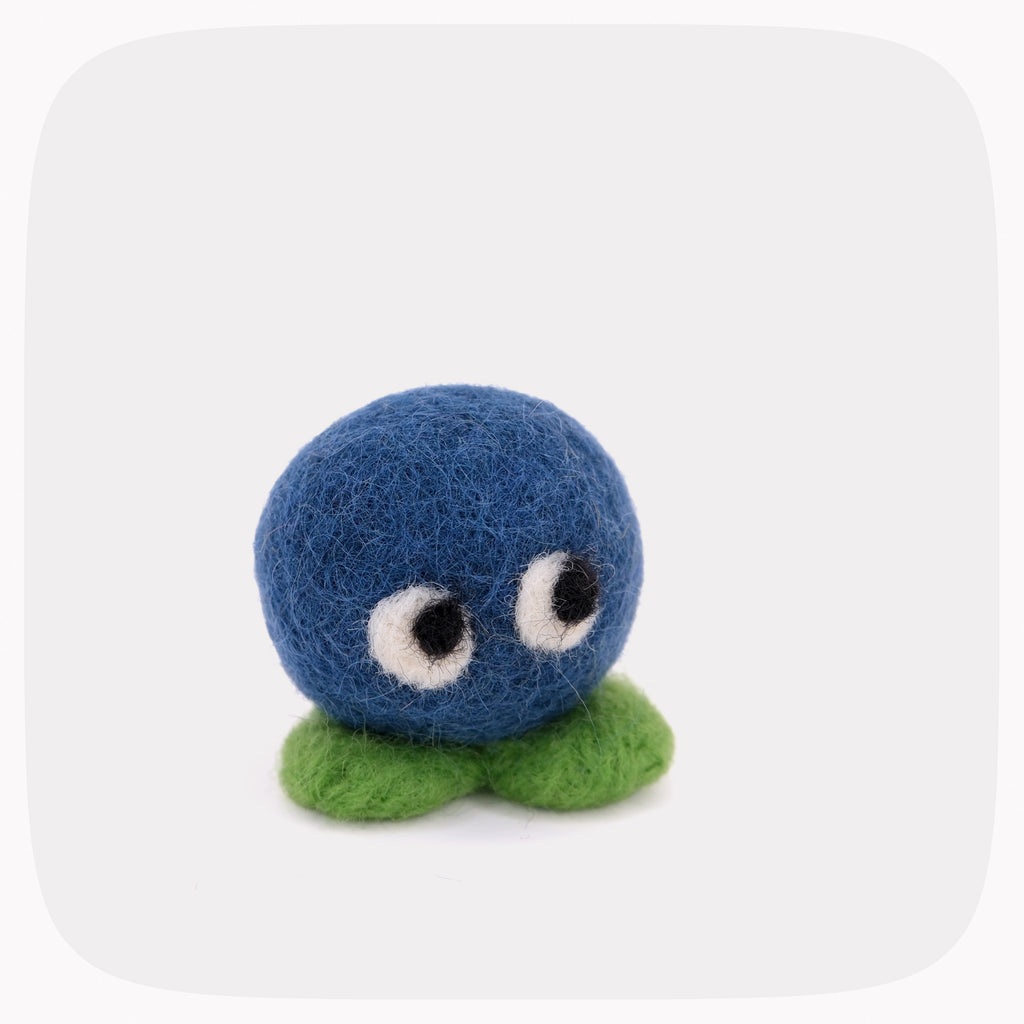 A small felted wool toy character with a round blue body, white eyes, and green feet, centered on a light gray rounded-square background.