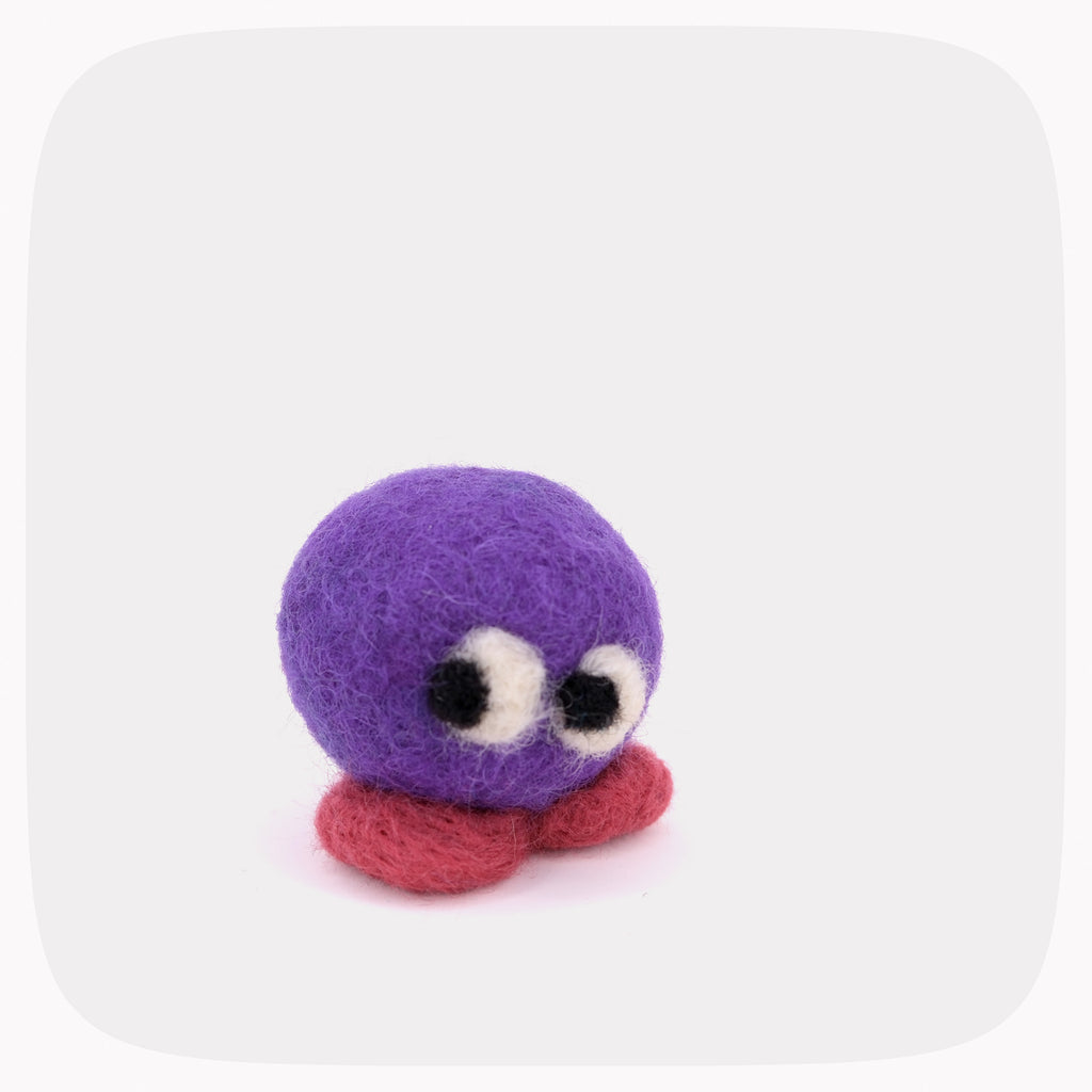 A small felted wool creature toy with a round purple head, large white and black eyes, and a red feet, centered on a light gray background with rounded corners.