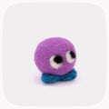 A small felted wool creature toy with a round purple head, large white and black eyes, and a blue textured base, centered on a light gray background with rounded corners.