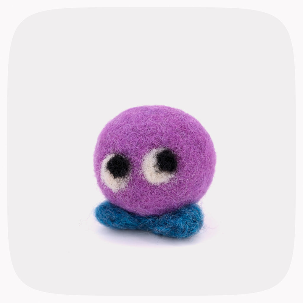 A small felted wool character toy with a round purple head, large black-and-white eyes, and a blue base, centered on a light gray background with rounded corners.