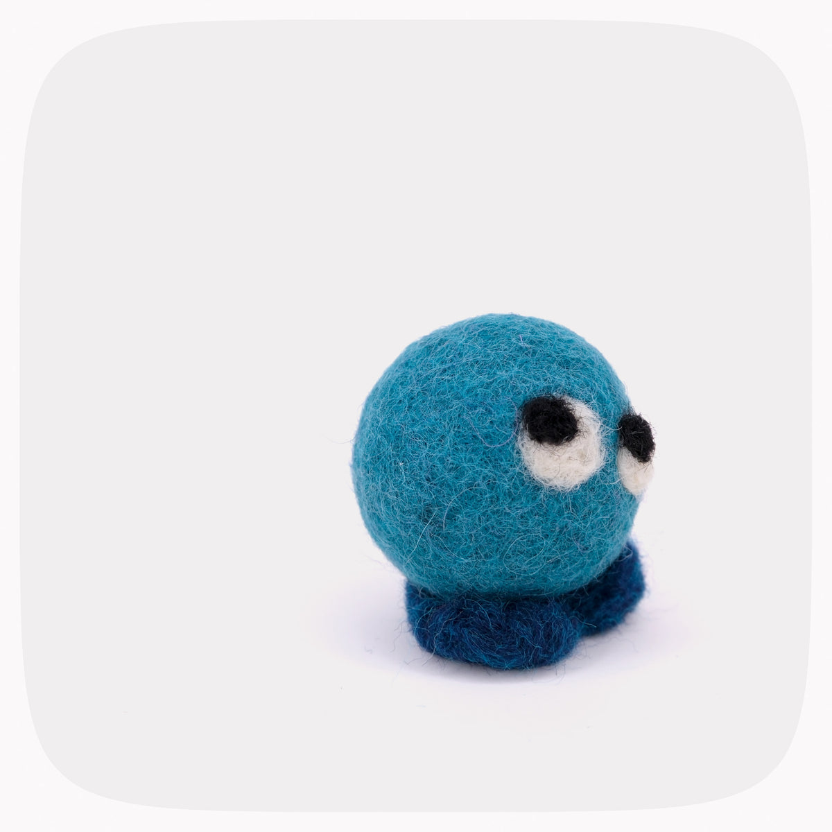 A small felted wool toy with a teal blue round body, dark blue feet, and large white eyes with black pupils, centered on a light gray background with rounded corners.