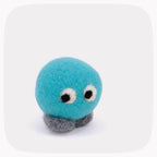 A small felt toy character with a round turquoise-blue head, gray feet, and large white-and-black eyes, photographed on a light gray background with rounded corners.