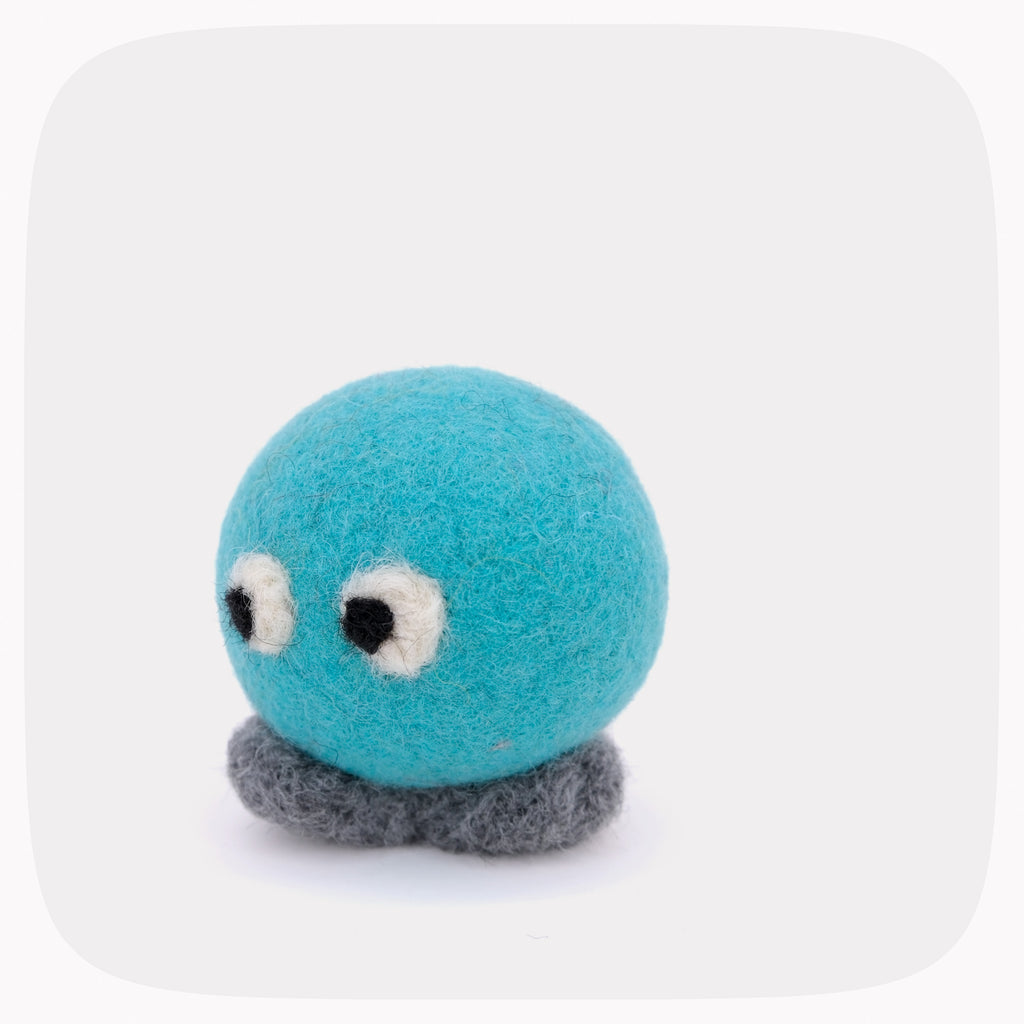 A small felt toy with a turquoise blue round head, white and black eyes, and a gray feet photographed on a light gray background.