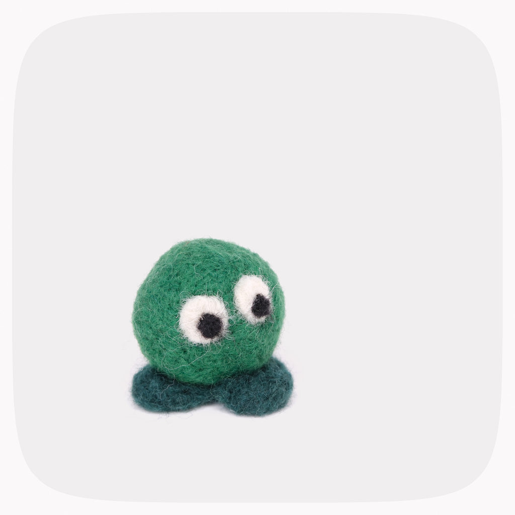 A small felted wool creature toy with a round green body, large white eyes with black pupils, and two dark teal feet, centered on a light gray rounded-square background.
