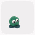 A small felted wool creature toy with a round green body, large white eyes with black pupils, and two dark teal feet, centered on a light gray rounded-square background.
