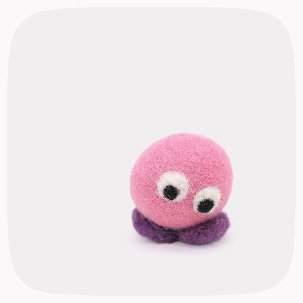 A small felt toy with a pink round body, purple feet and large googly-style eyes, photographed on a light gray background with rounded corners.